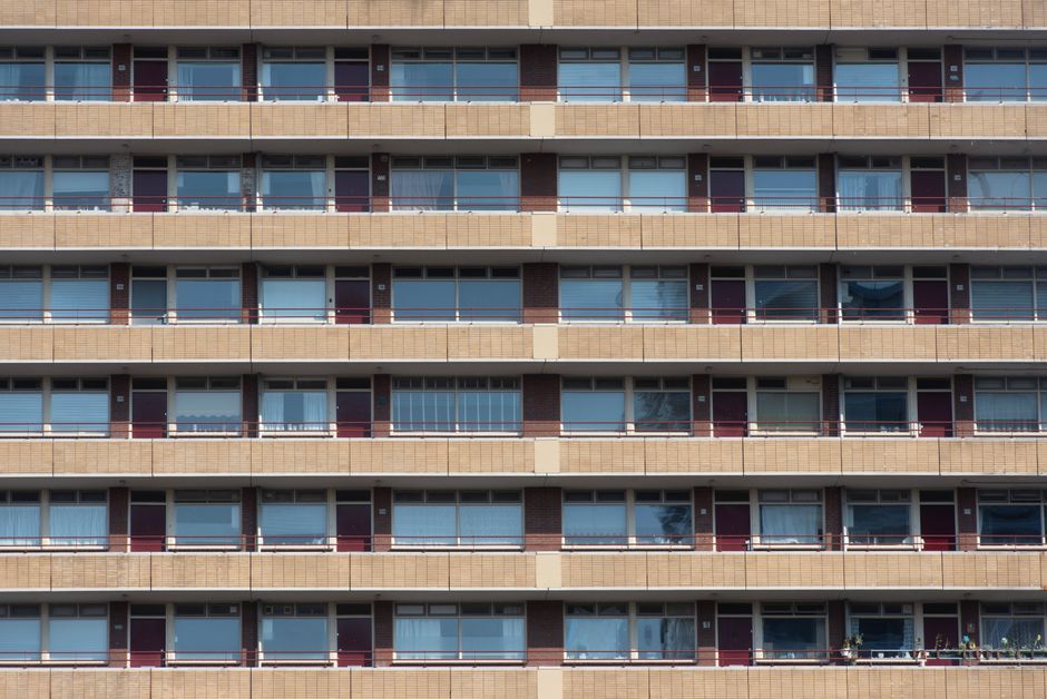 A close-up view of a multi-story residential building's exterior featuring rows of large glass windows with white frames, set into a brick facade with tan-colored horizontal bands. The windows reflect