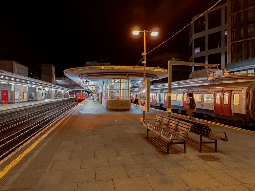 A train arriving at Clapham Junction station platform, showcasing stainless steel train cars with a glossy, clean surface reflecting sunlight. The platform has a textured yellow safety line along the